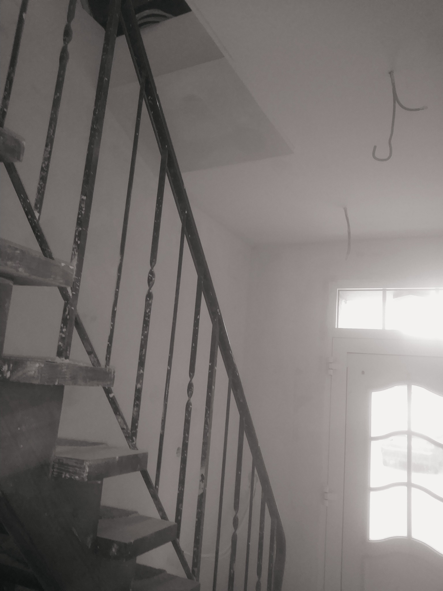 Klatka schodowa w trakcie prac wykończeniowych, widoczne drewniane schody z metalową balustradą, ślady farby na stopniach i balustradzie, wystające przewody z sufitu, drzwi z szybami w tle.