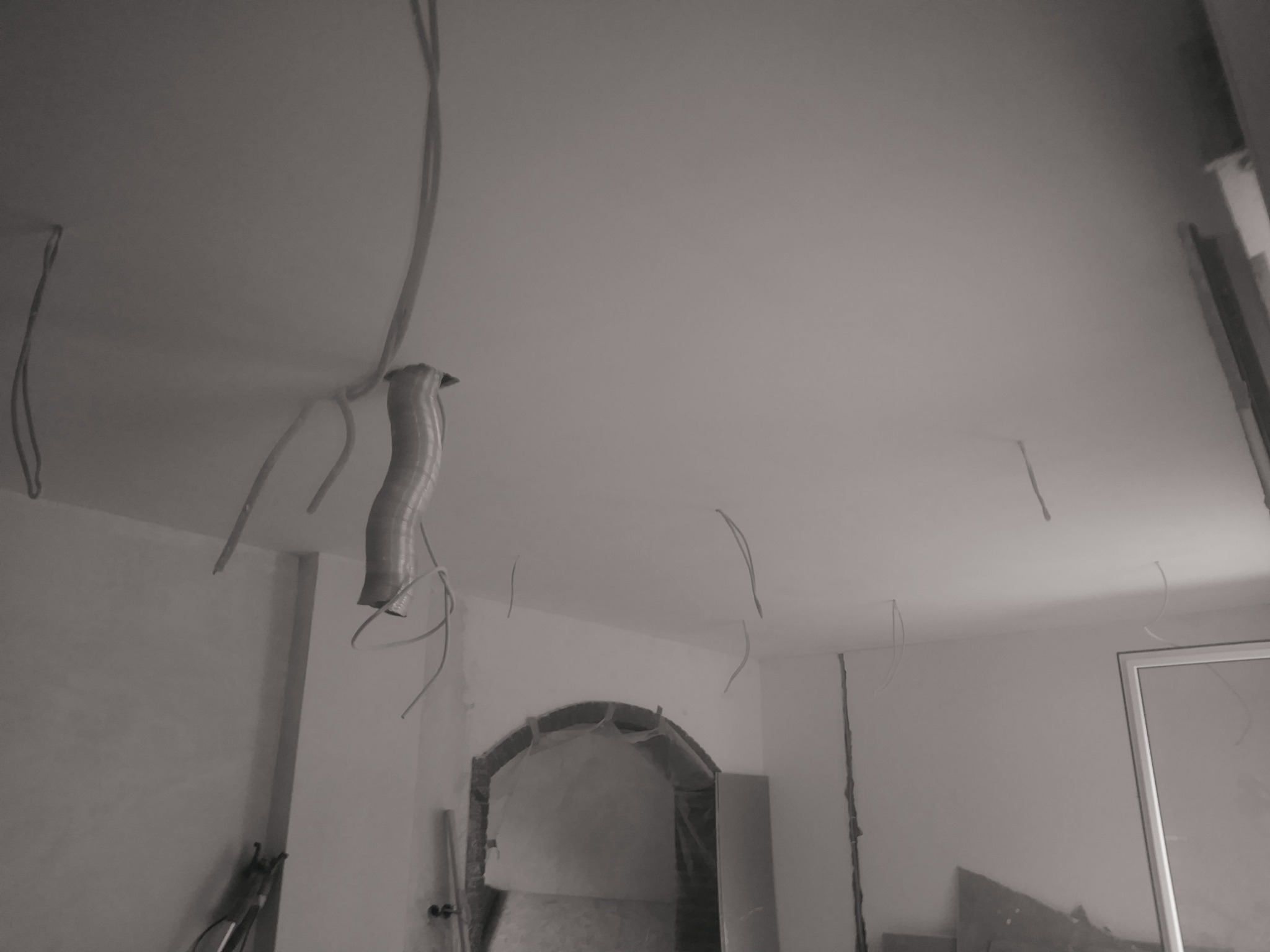 Wnętrze w trakcie prac: widoczne kable zwisające z sufitu, przygotowany otwór drzwiowy z folią ochronną i ściany w szarym odcieniu przed wykończeniem.