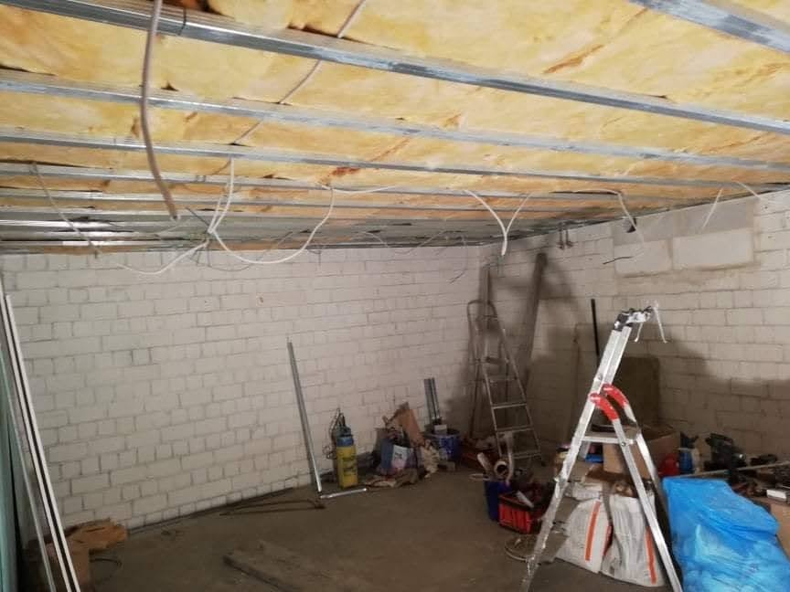 Ocieplenie sufitu wełną mineralną w pomieszczeniu z białej cegły, widoczne metalowe profile, przewody elektryczne, drabiny i narzędzia budowlane.