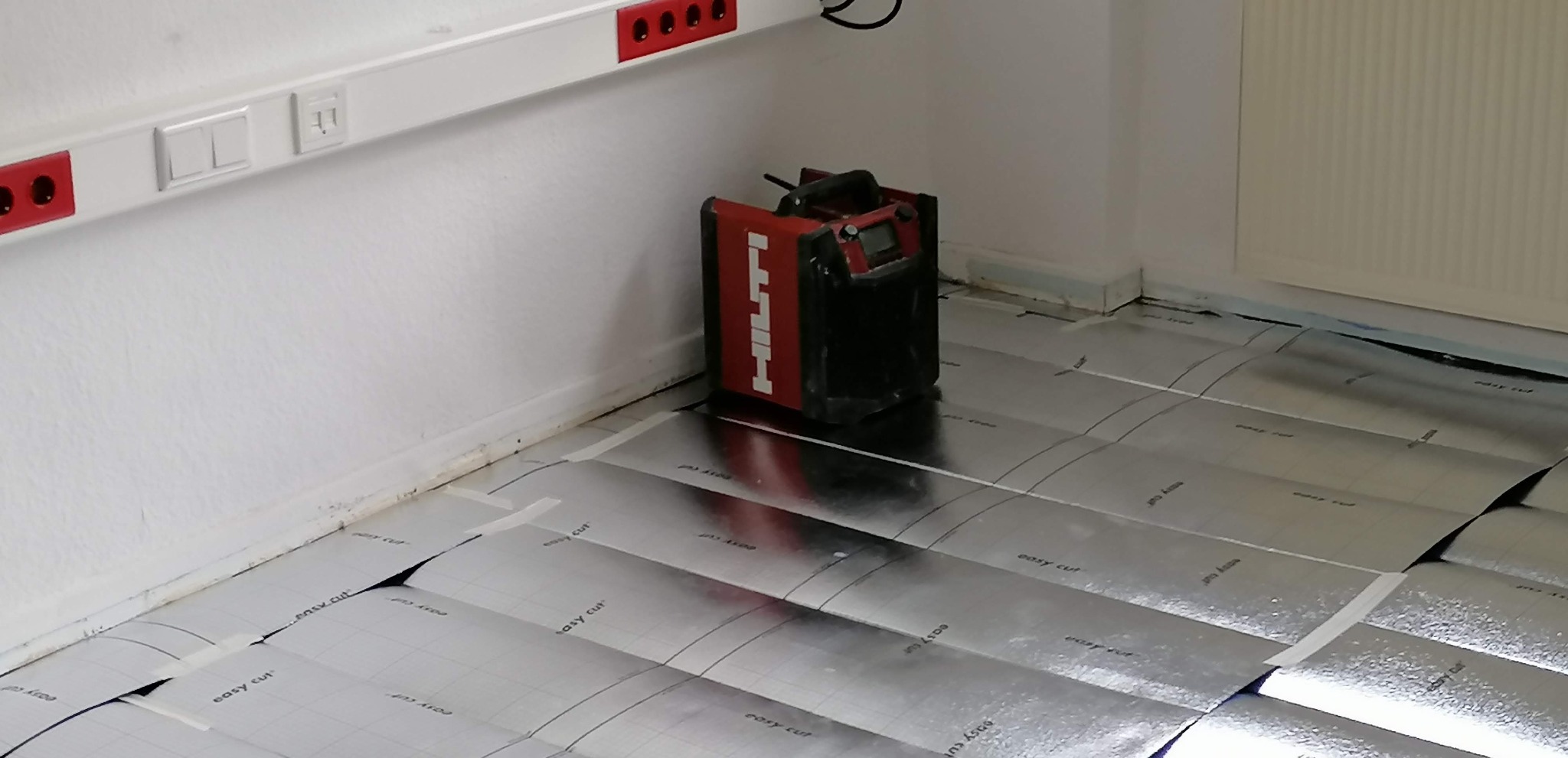 Przygotowanie podłoża pod ogrzewanie podłogowe: folia aluminiowa z nadrukiem 'easy cut' rozłożona na podłodze, obok poziomica laserowa Hilti, widoczne listwy przypodłogowe i gniazdka elektryczne.