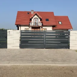 Nowoczesna brama wjazdowa i furtka z poziomymi panelami w kolorze grafitowym, osadzone w białych, modułowych słupkach betonowych, na tle domu z czerwoną dachówką i brukowanym podjazdem.