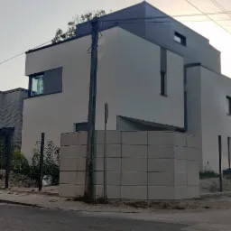 Nowoczesny dom z ogrodzeniem z betonowych paneli w trakcie montażu, widoczne słupki i niedokończone fragmenty, poranne słońce