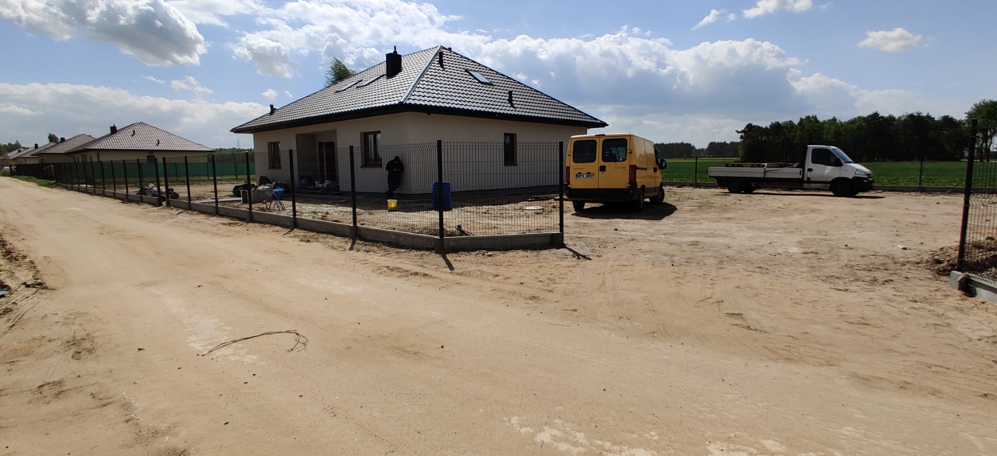 Nowo postawiony dom jednorodzinny z czarną, metalową siatką ogrodzeniową na betonowej podmurówce, z żółtym busem i białym samochodem dostawczym na posesji.