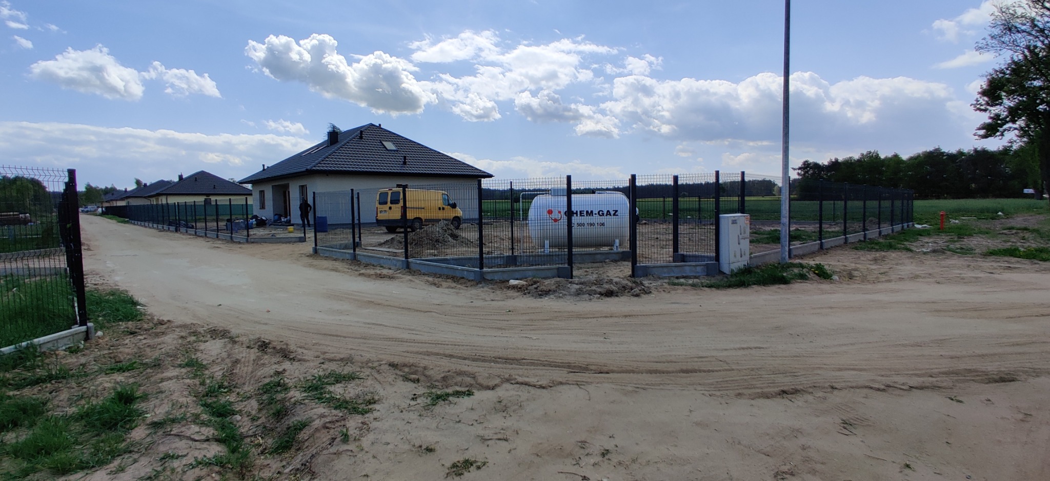 Montaż czarnego ogrodzenia panelowego z podmurówką betonową na nowym osiedlu domów jednorodzinnych, widoczny żółty samochód dostawczy i zbiornik gazu.