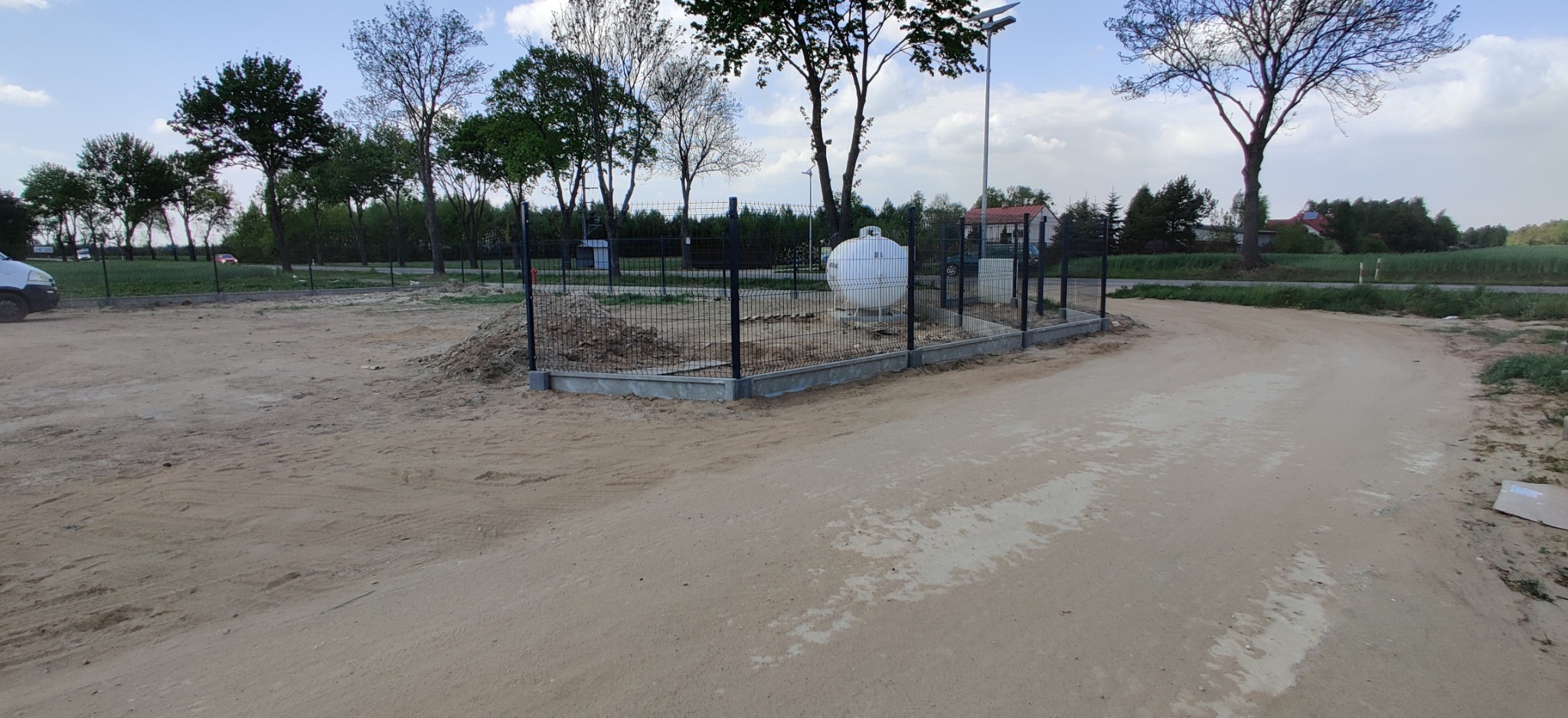 Montaż czarnego ogrodzenia panelowego wokół zbiornika gazu na posesji, widoczne słupy i betonowa podmurówka, w tle drzewa i budynki.