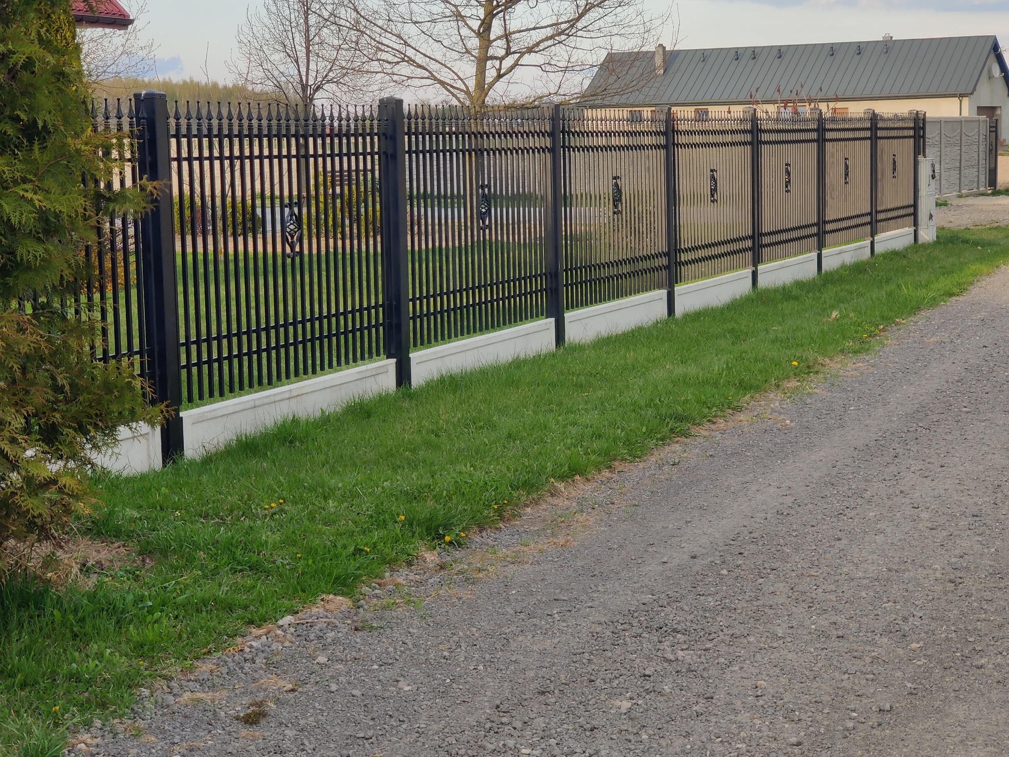 Czarny, metalowy płot z ozdobnymi elementami, osadzony na betonowej podmurówce, widoczny z perspektywy gruntu, z domem w tle.