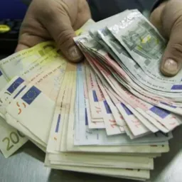 Dłonie trzymające wachlarz banknotów euro o różnych nominałach (5, 10, 200), ułożonych na metalowej powierzchni