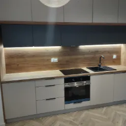 Nowoczesna kuchnia z beżowymi szafkami, drewnianym blatem i czarnymi elementami, oświetlona lampą sufitową i listwą LED, z widocznym odkurzaczem na parkiecie w jodełkę.