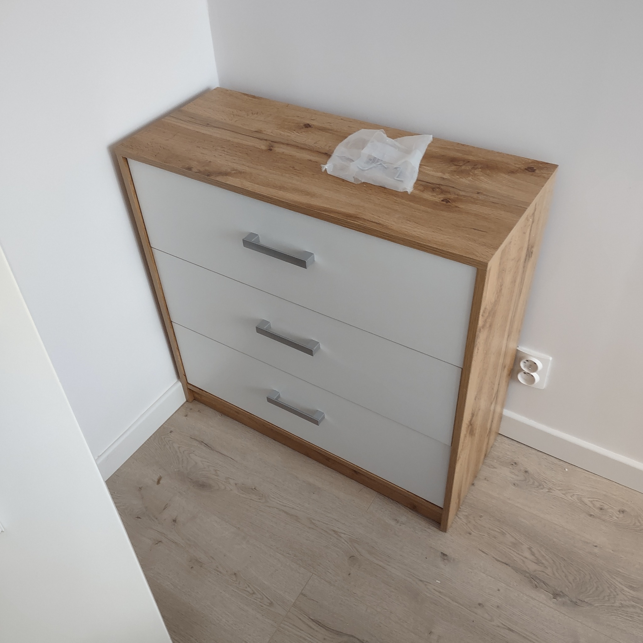 Komoda z trzema szufladami w kolorze dębu i bieli, z metalowymi uchwytami, stoi przy białej ścianie z widocznym gniazdkiem elektrycznym, na blacie leży woreczek z akcesoriami montażowymi.