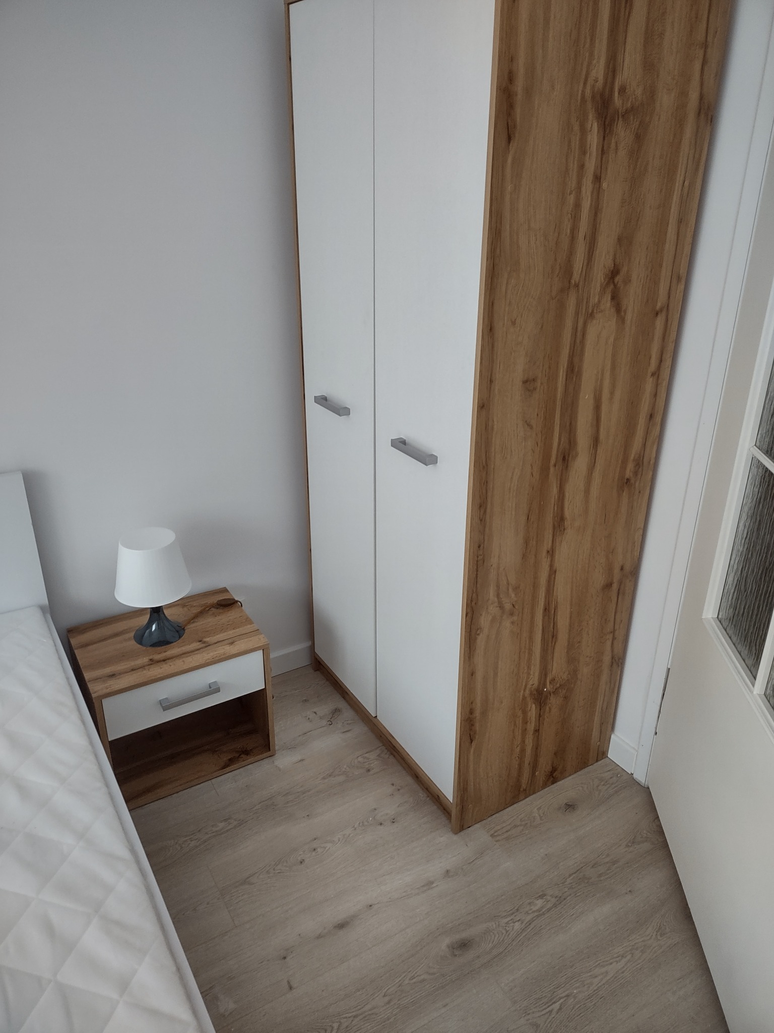 Dwudrzwiowa szafa z białymi frontami i bokami w kolorze drewna, stojąca obok szafki nocnej z lampką, widok z góry.