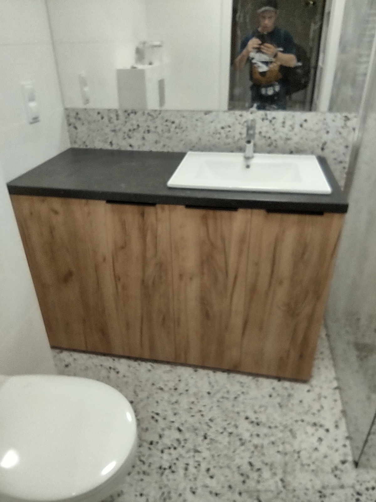 Umywalka z szafką z drewnopodobnymi frontami i czarnym blatem, widoczny fragment toalety, odbicie osoby robiącej zdjęcie w lustrze.