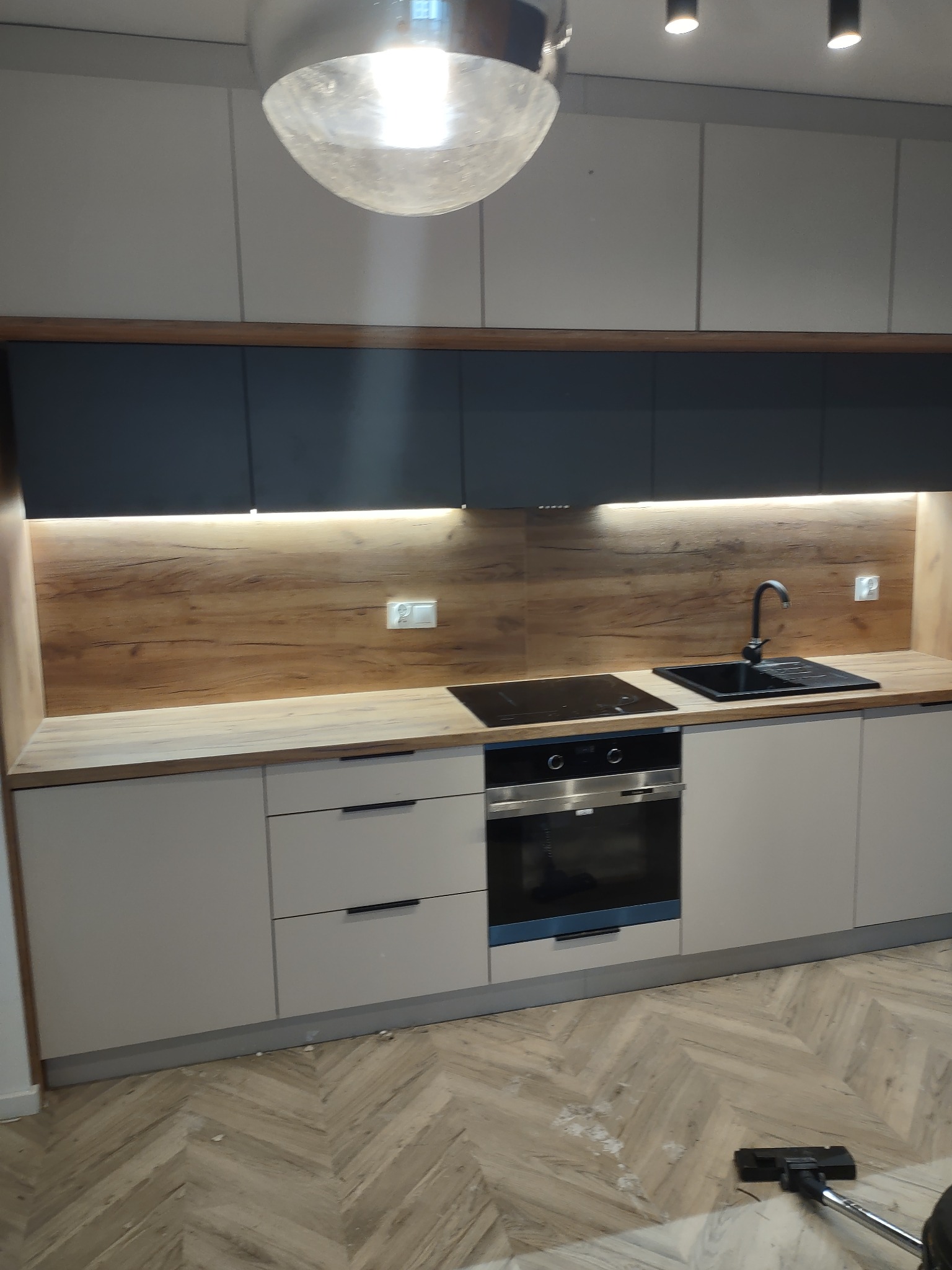 Nowoczesna kuchnia z beżowymi szafkami, drewnianym blatem i czarnymi elementami, oświetlona lampą sufitową i listwą LED, z widocznym odkurzaczem na parkiecie w jodełkę.