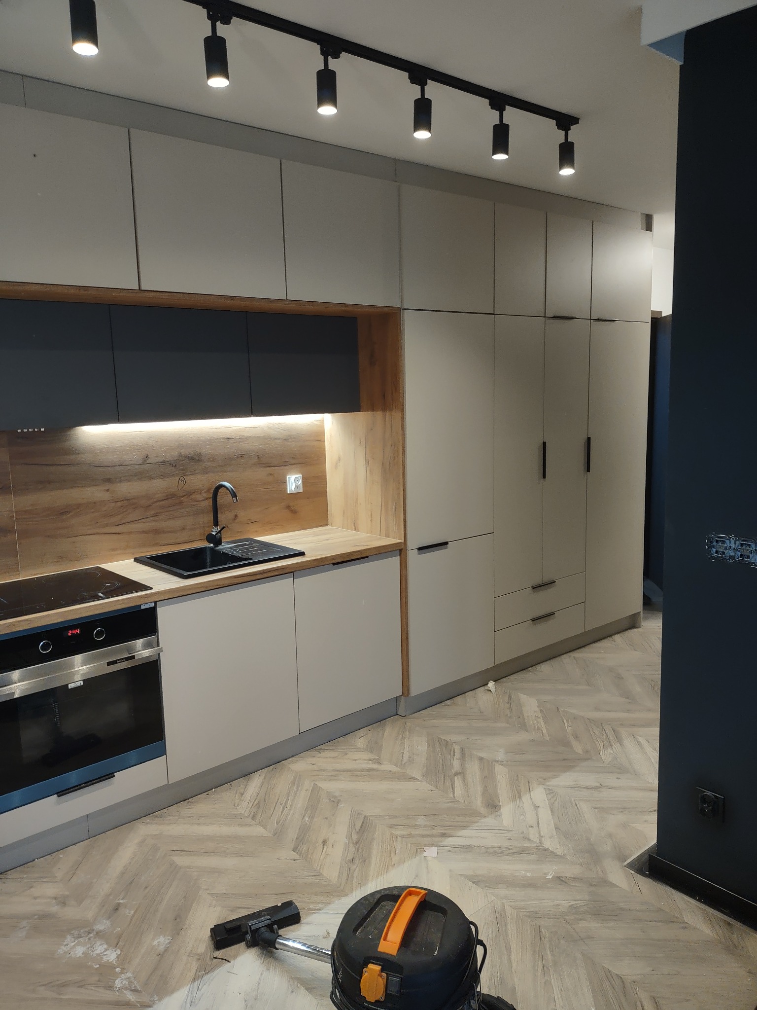 Nowoczesna kuchnia z beżowymi szafkami, drewnianym blatem i czarnym zlewem, po montażu, z widocznymi śladami po pracy i odkurzaczem na podłodze w jodełkę.