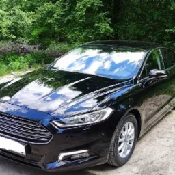 Czarny Ford Mondeo Hybrid zaparkowany na piaszczystej drodze z zielonymi drzewami w tle, z widocznymi odbiciami chmur na karoserii.