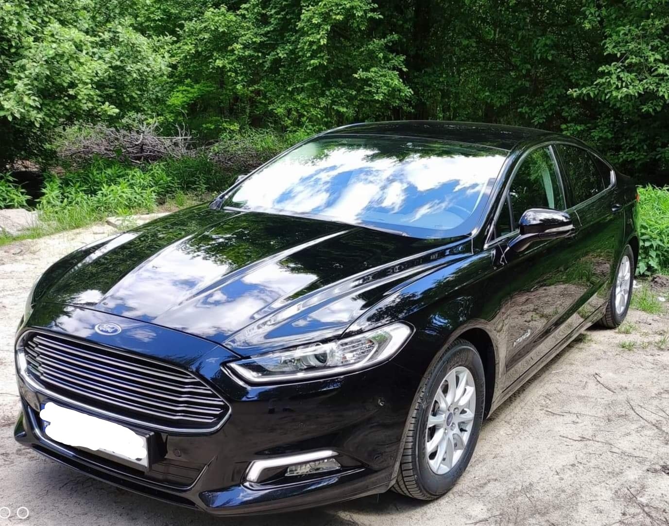 Czarny Ford Mondeo Hybrid zaparkowany na piaszczystej drodze z zielonymi drzewami w tle, z widocznymi odbiciami chmur na karoserii.