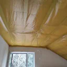 Żółta folia paroizolacyjna szczelnie pokrywająca krokwie poddasza, widoczne okno dachowe i fragment ściany przed wykończeniem.