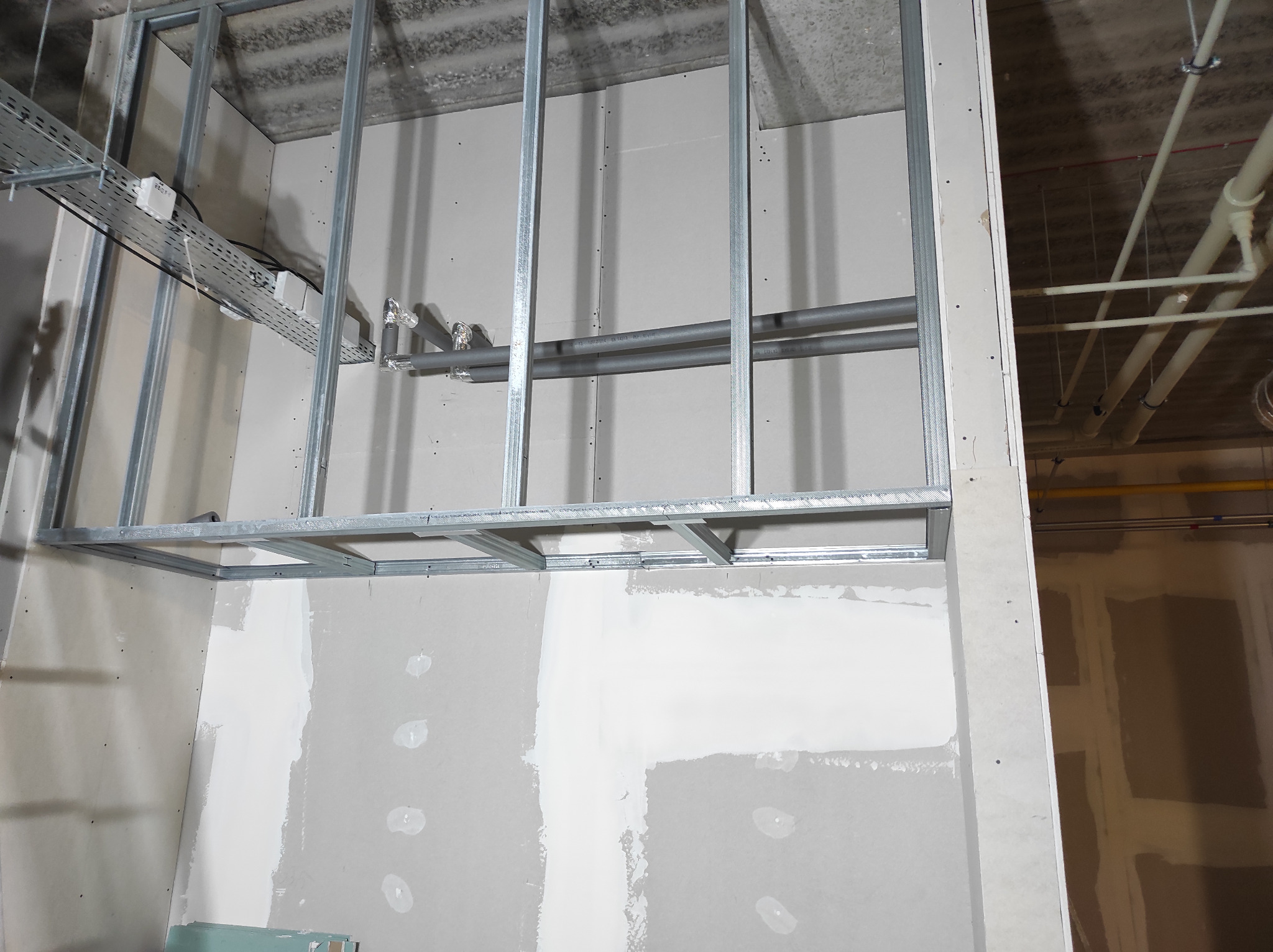 Niedokończona ściana z metalowym szkieletem, częściowo pokryta płytami, widoczne instalacje pod sufitem.
