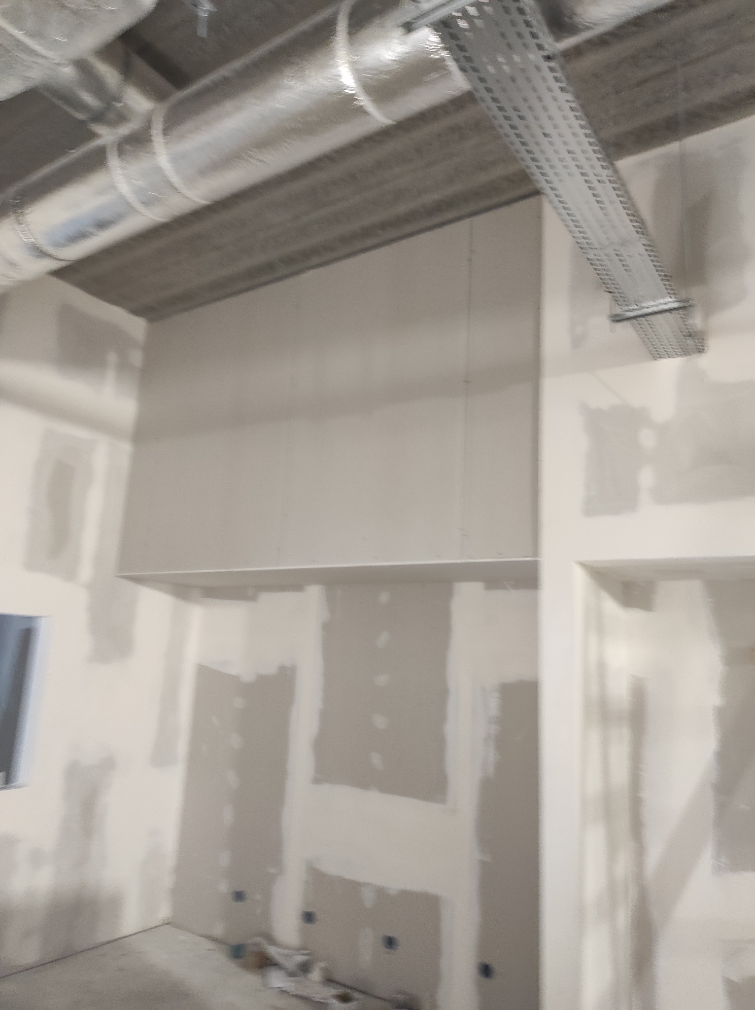 Montaż płyty karton gips na ścianach i suficie w surowym wnętrzu, widoczne rury wentylacyjne i koryto kablowe pod sufitem.