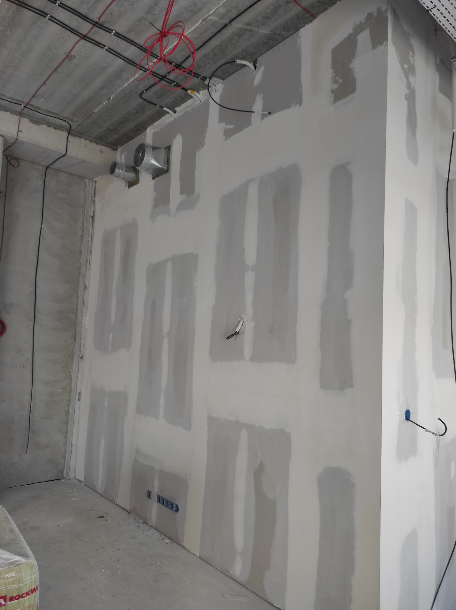 Wnętrze w trakcie wykańczania: ściany z zamontowanymi i częściowo zaszpachlowanymi płytami kartonowo-gipsowymi, widoczne przewody elektryczne pod sufitem oraz wentylacja.