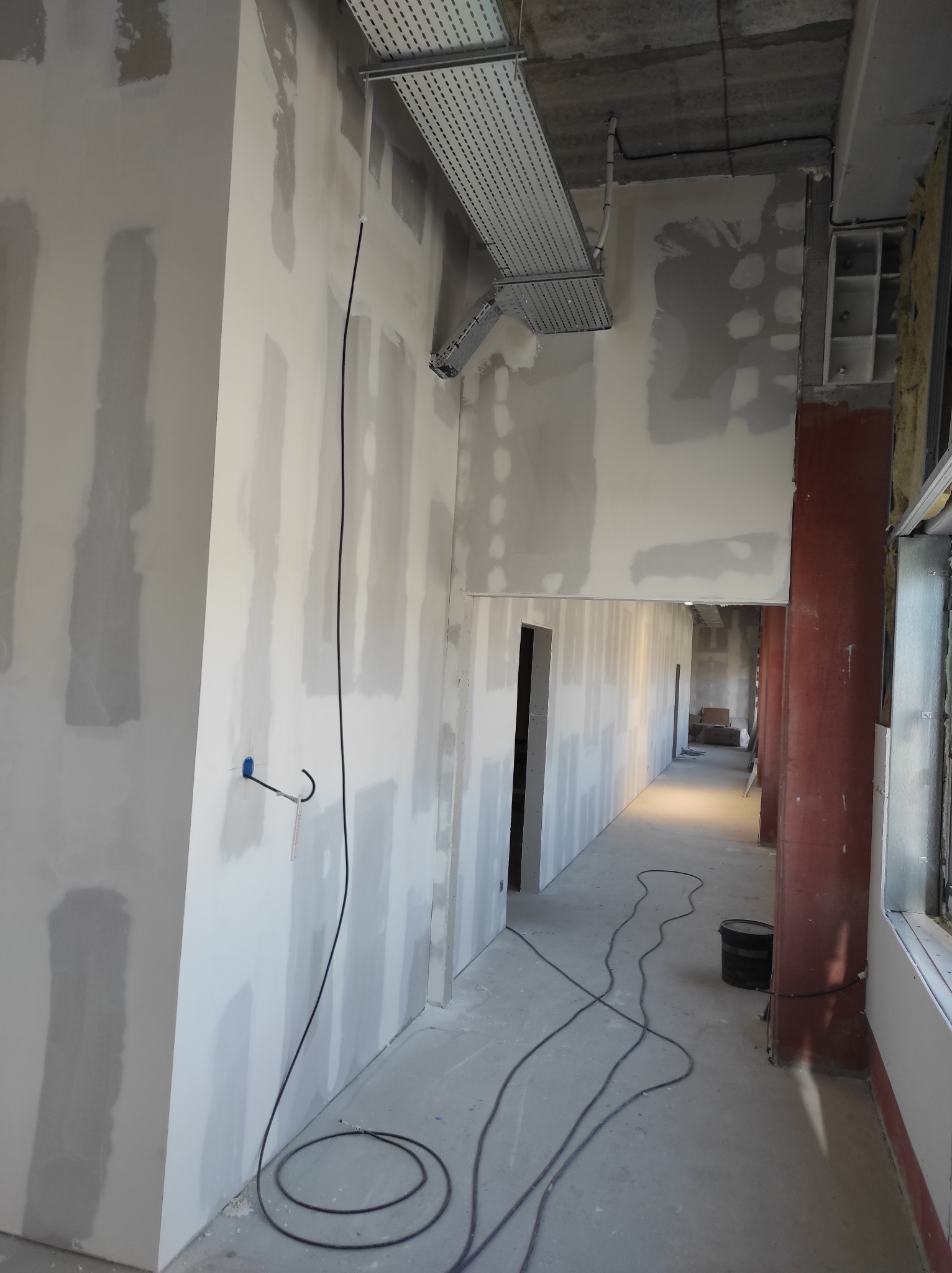 Korytarz w trakcie wykańczania, ściany pokryte szpachlą na łączeniach płyt, widoczne kable i puszka instalacyjna, sufit z elementami instalacji wentylacyjnej.