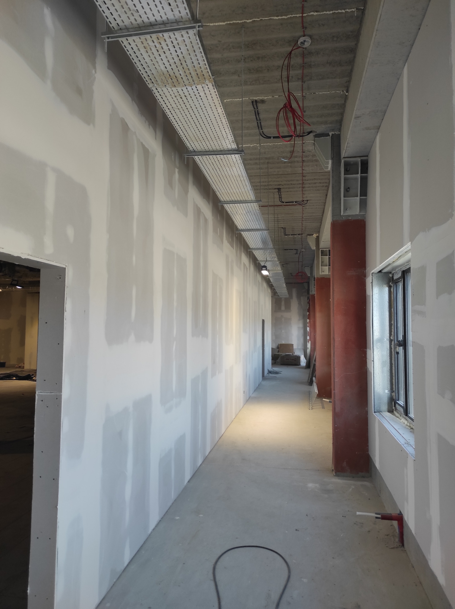 Korytarz w trakcie wykańczania, ściany pokryte płytami gipsowo-kartonowymi z widocznymi łączeniami przed szpachlowaniem, instalacje elektryczne pod sufitem, widoczne czerwone kolumny i okno.