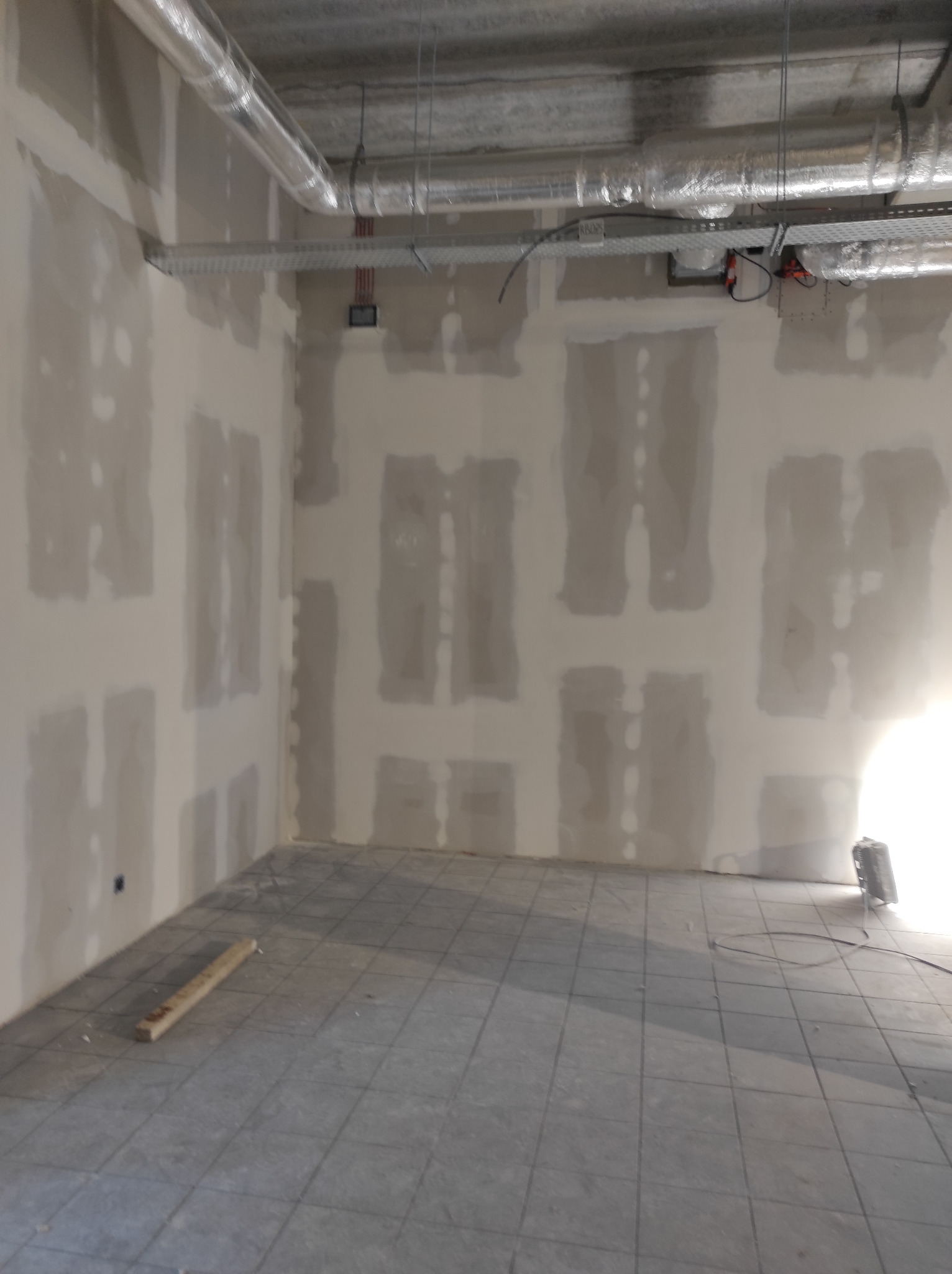 Wnętrze w trakcie wykańczania: ściany z widocznymi łączeniami płyt, szare płytki na podłodze, instalacje wentylacyjne pod sufitem. Na podłodze leży drewniana listwa.