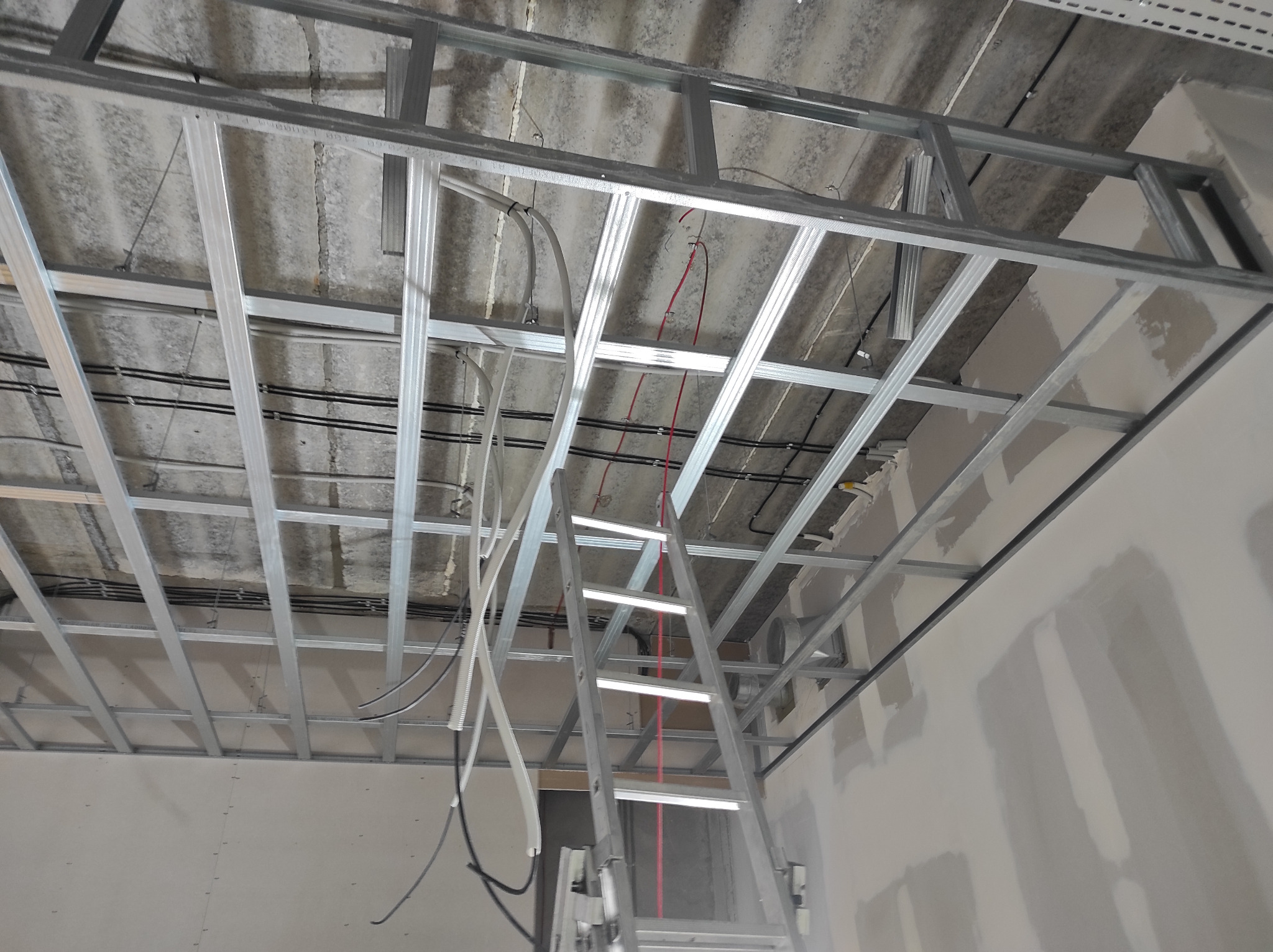 Metalowa konstrukcja sufitu podwieszanego z widocznymi przewodami i schodami drabinkowymi w pomieszczeniu w trakcie prac wykończeniowych.