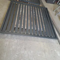 Dwa metalowe panele ogrodzeniowe w kolorze szarym, ułożone na betonowej podłodze, prawdopodobnie w warsztacie, z widocznym sprzętem i materiałami w tle.