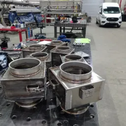 Metalowe, spawane konstrukcje skrzynkowe z okrągłymi otworami na górze, ułożone na perforowanym stole spawalniczym w hali produkcyjnej, z widocznym samochodem dostawczym w tle.