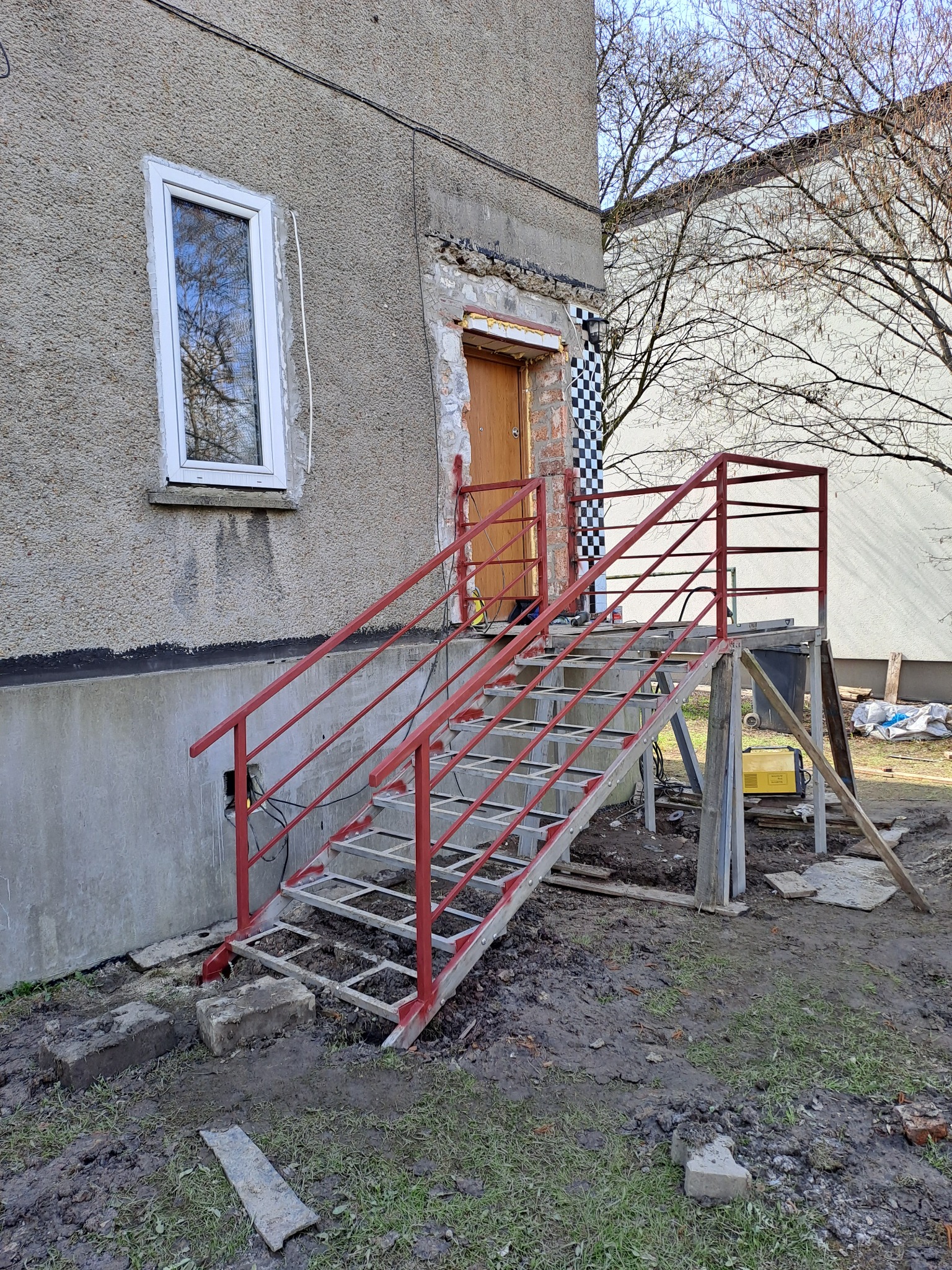 Zewnętrzne schody metalowe z czerwoną balustradą, w trakcie montażu przy szarym budynku z widocznymi śladami prac budowlanych i ziemistym podłożem.