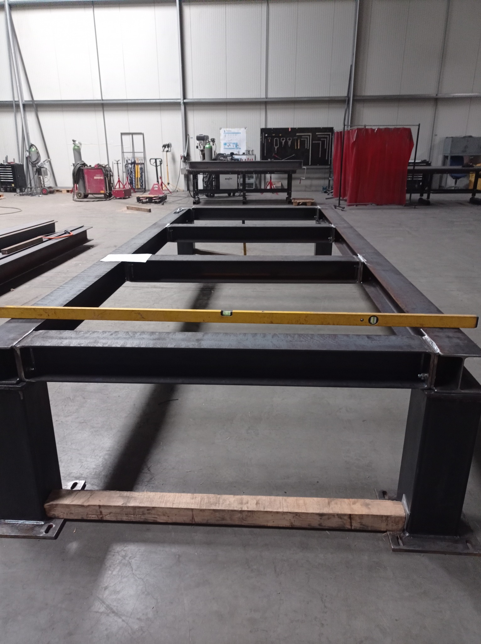 Konstrukcja stalowa w hali produkcyjnej, z poziomnicą na górnej ramie, drewniane podkładki pod nogami, widoczne narzędzia i sprzęt spawalniczy w tle.