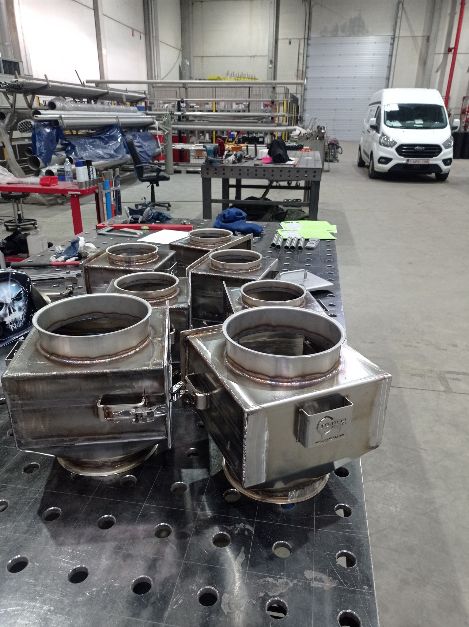 Metalowe, spawane konstrukcje skrzynkowe z okrągłymi otworami na górze, ułożone na perforowanym stole spawalniczym w hali produkcyjnej, z widocznym samochodem dostawczym w tle.