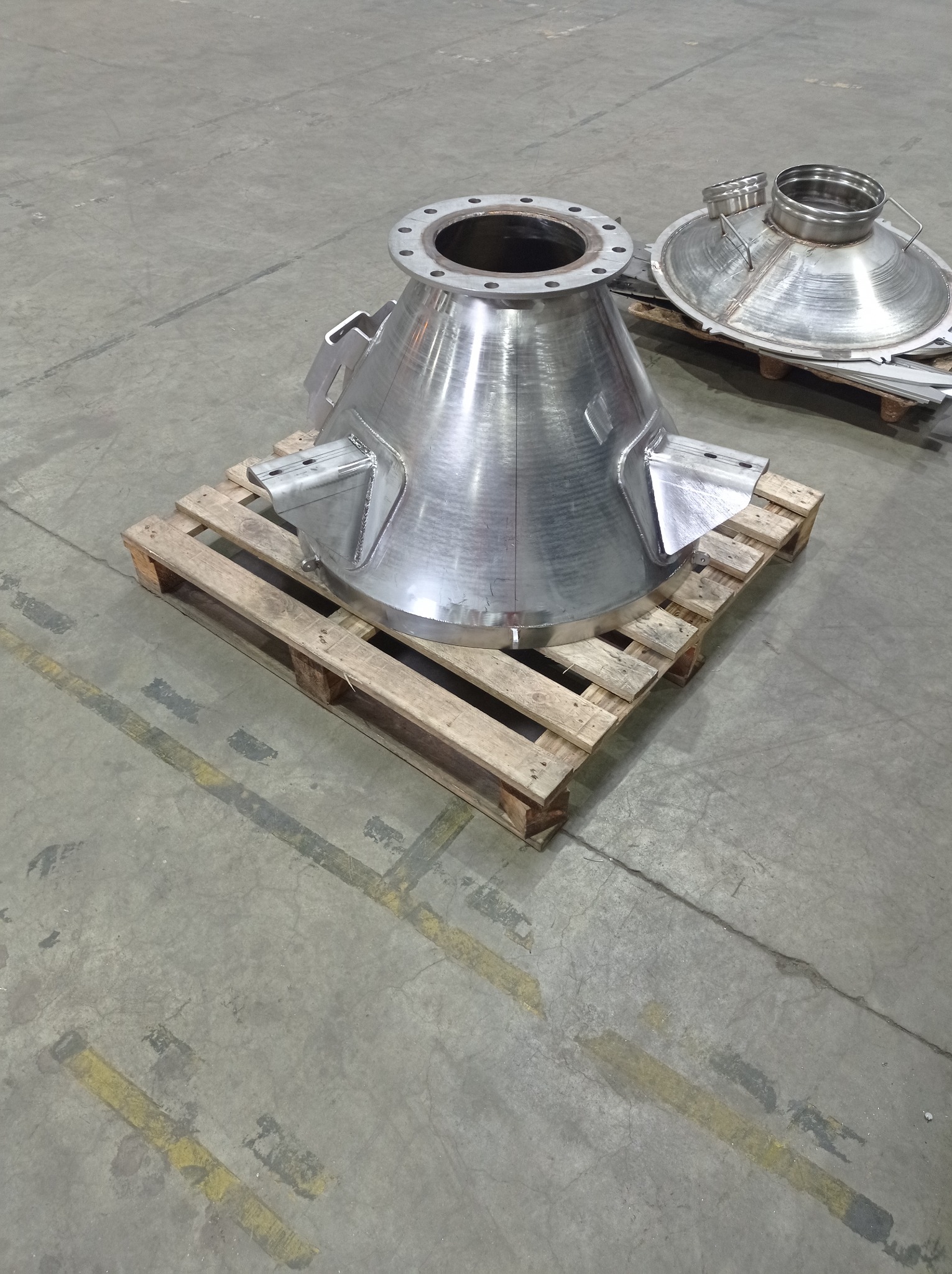 Stożkowy element ze stali nierdzewnej na palecie, z widocznymi spawami i kołnierzem z otworami, obok znajduje się pokrywa.