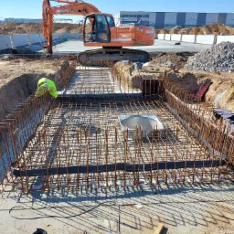 Budowa fundament&oacute;w: Zbrojenie klatki z pręt&oacute;w zbrojeniowych, widoczny pracownik w ż&oacute;łtej kamizelce, koparka Doosan w tle, teren budowy.