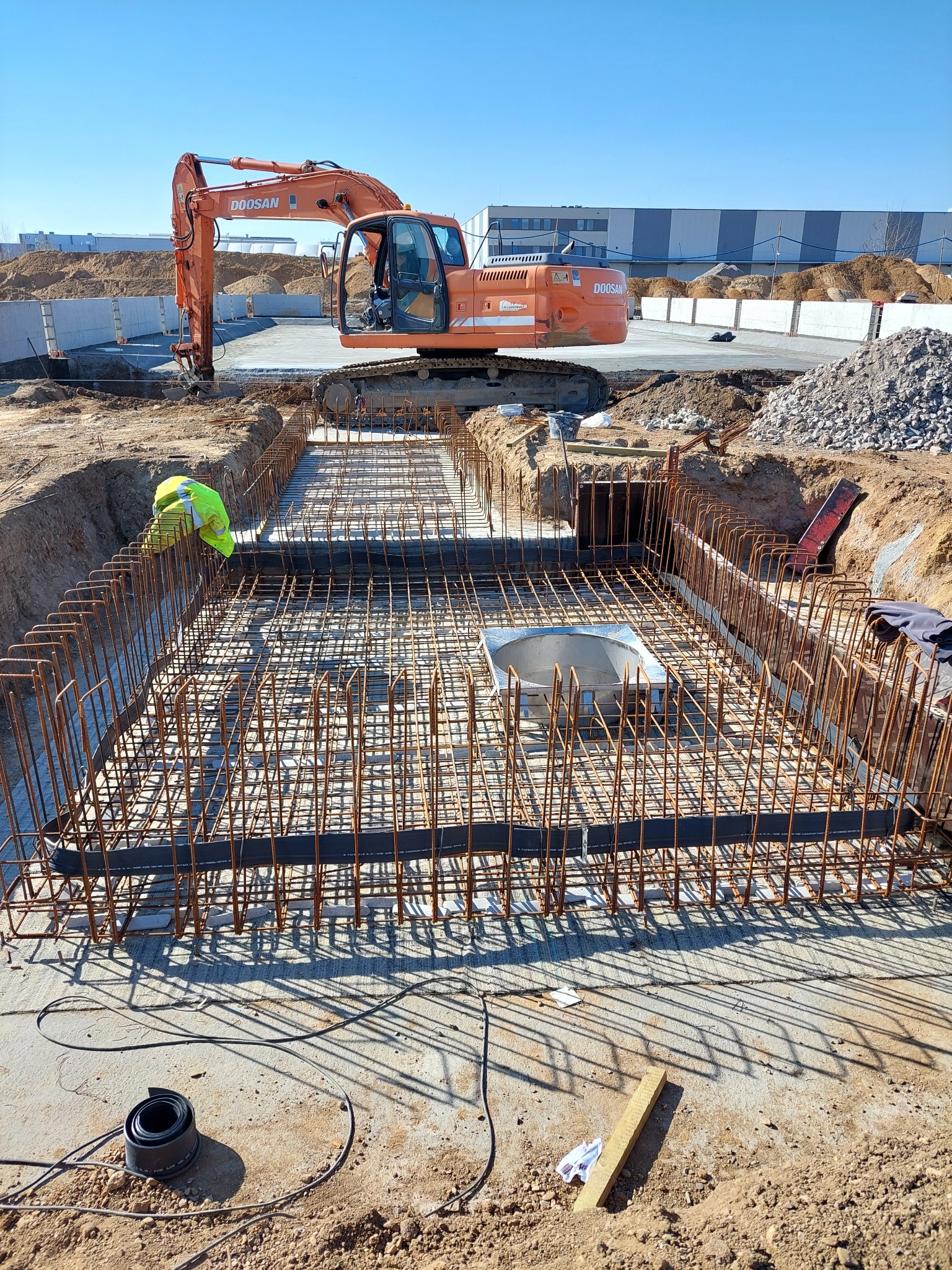Budowa fundamentów: Zbrojenie klatki z prętów zbrojeniowych, widoczny pracownik w żółtej kamizelce, koparka Doosan w tle, teren budowy.