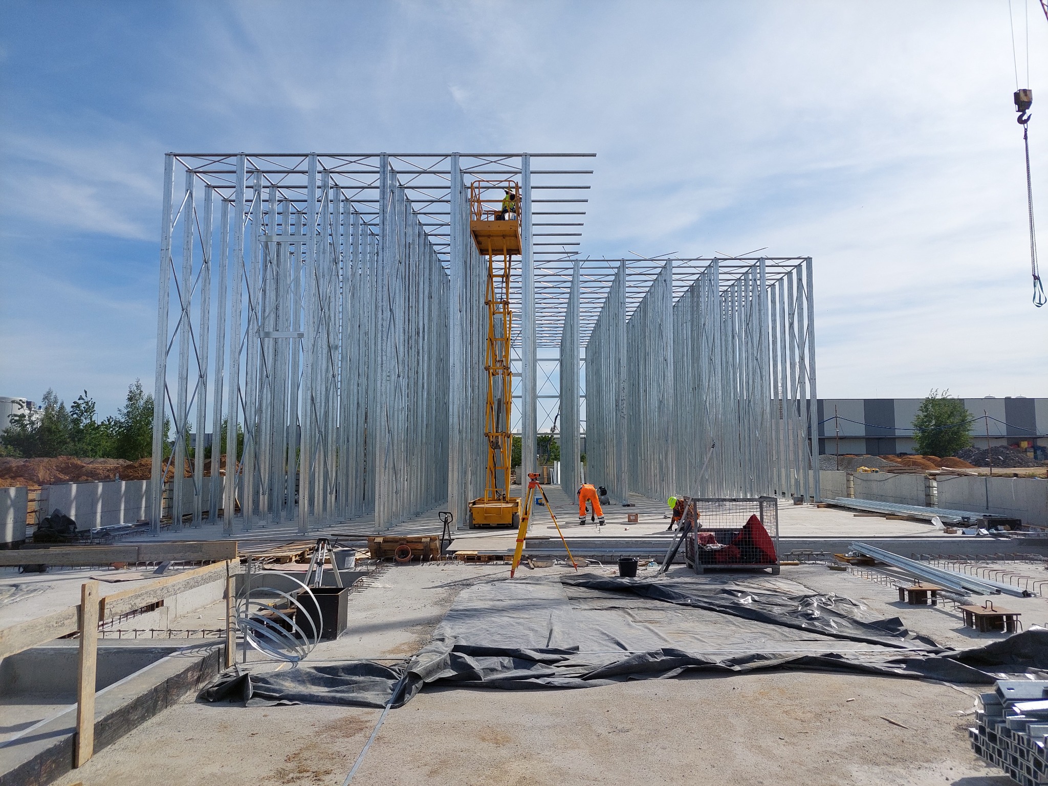 Stalowa konstrukcja budynku w trakcie montażu, pracownicy w odblaskowych ubraniach, żółty podnośnik koszowy i niwelator optyczny na placu budowy przy słonecznej pogodzie.