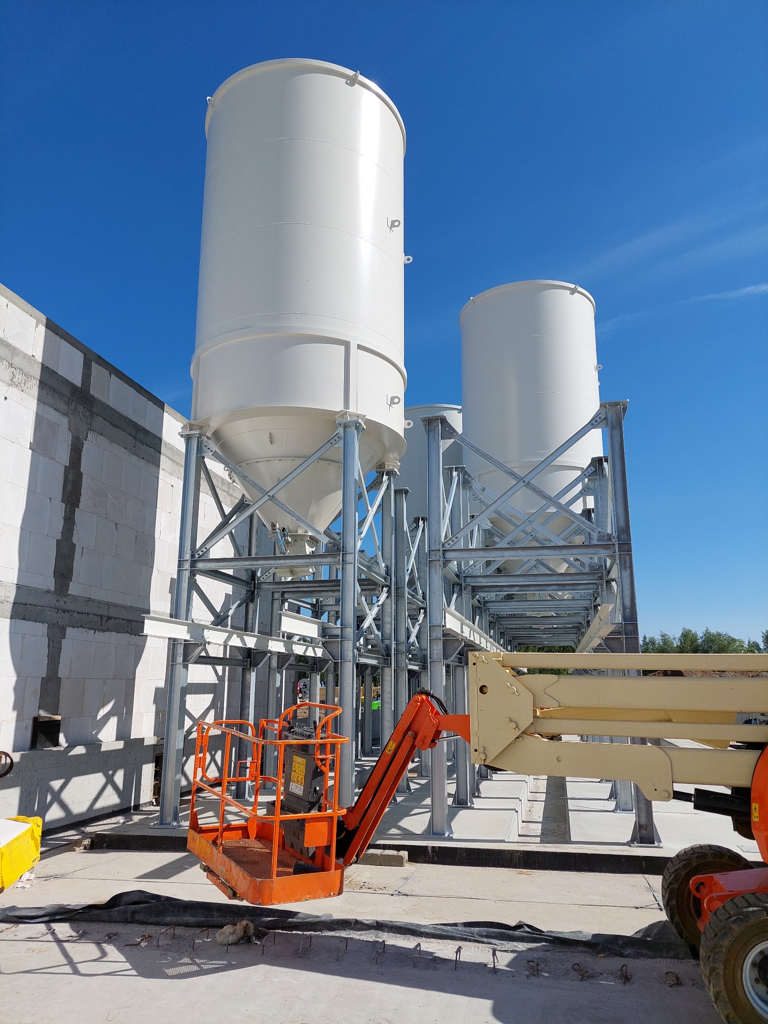 Dwa białe silosy na stalowych konstrukcjach, widoczne z perspektywy podnośnika koszowego, z fragmentem muru z betonu komórkowego w tle i błękitnym niebem.