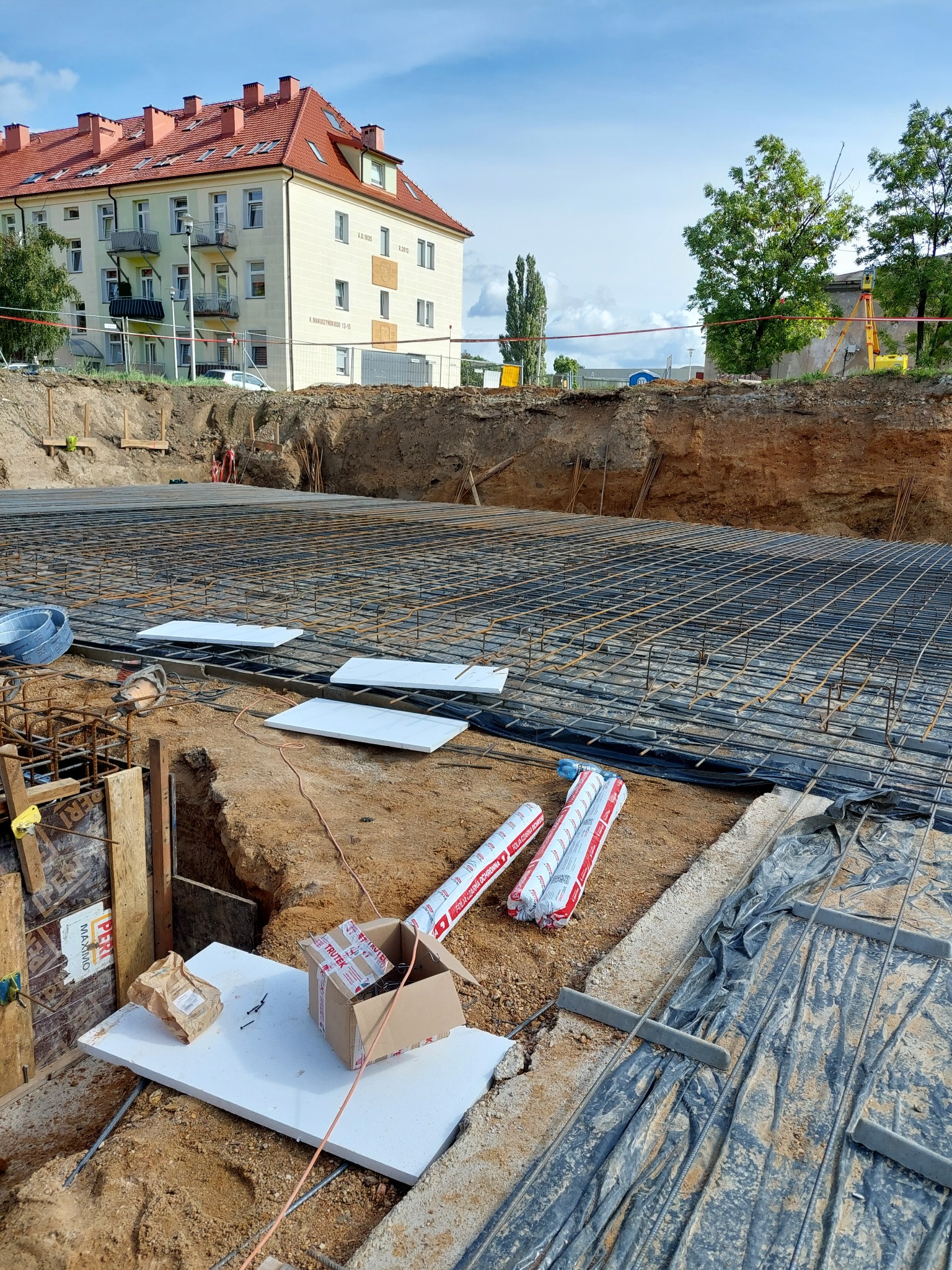 Przygotowanie zbrojenia fundamentów pod budynek wielorodzinny, widoczne siatki zbrojeniowe, folia, styropian, materiały izolacyjne w tubach, wykop ziemny i budynek mieszkalny w tle.