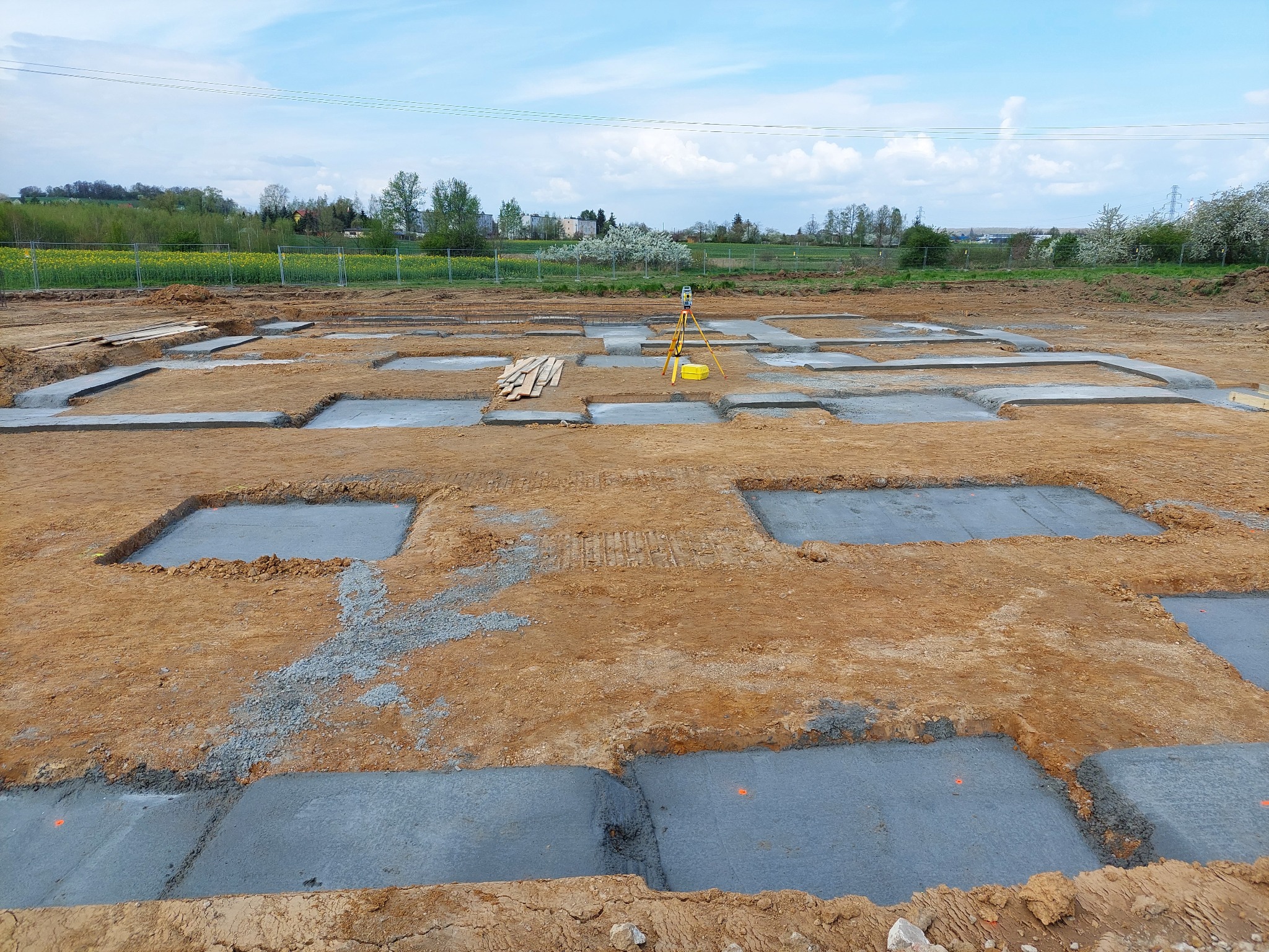 Wytyczone fundamenty pod budynek z użyciem niwelatora optycznego, widoczne deski szalunkowe i krajobraz z kwitnącymi drzewami w tle.