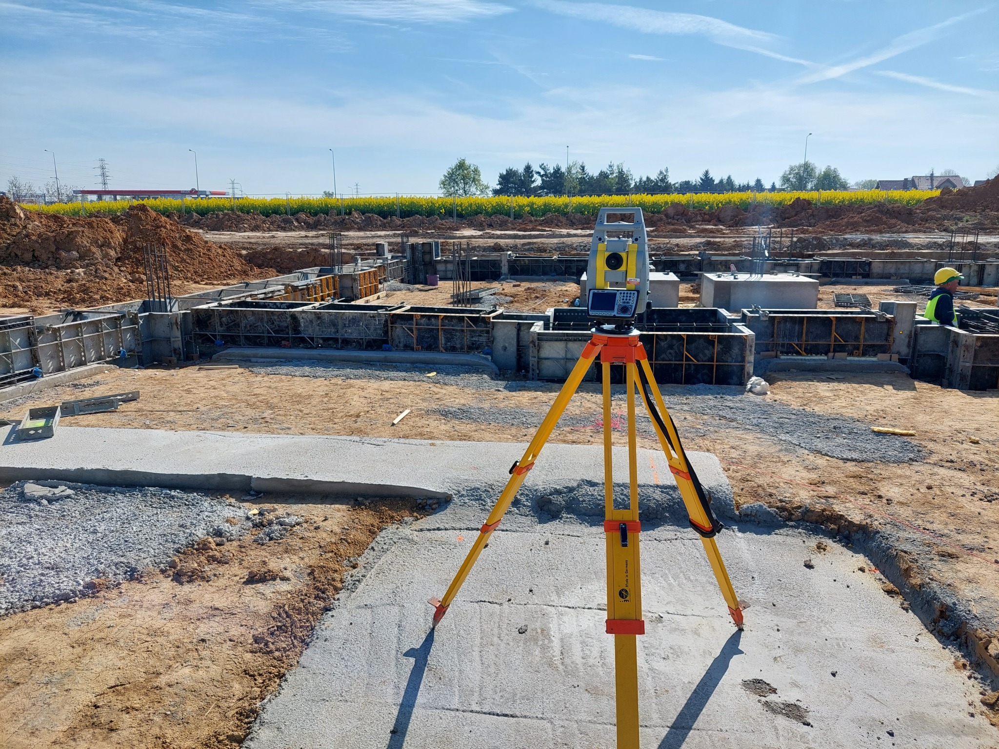 Żółty teodolit ustawiony na betonowej płycie fundamentowej w trakcie budowy, w tle widoczne żółte pola rzepaku i elementy konstrukcyjne budynku.