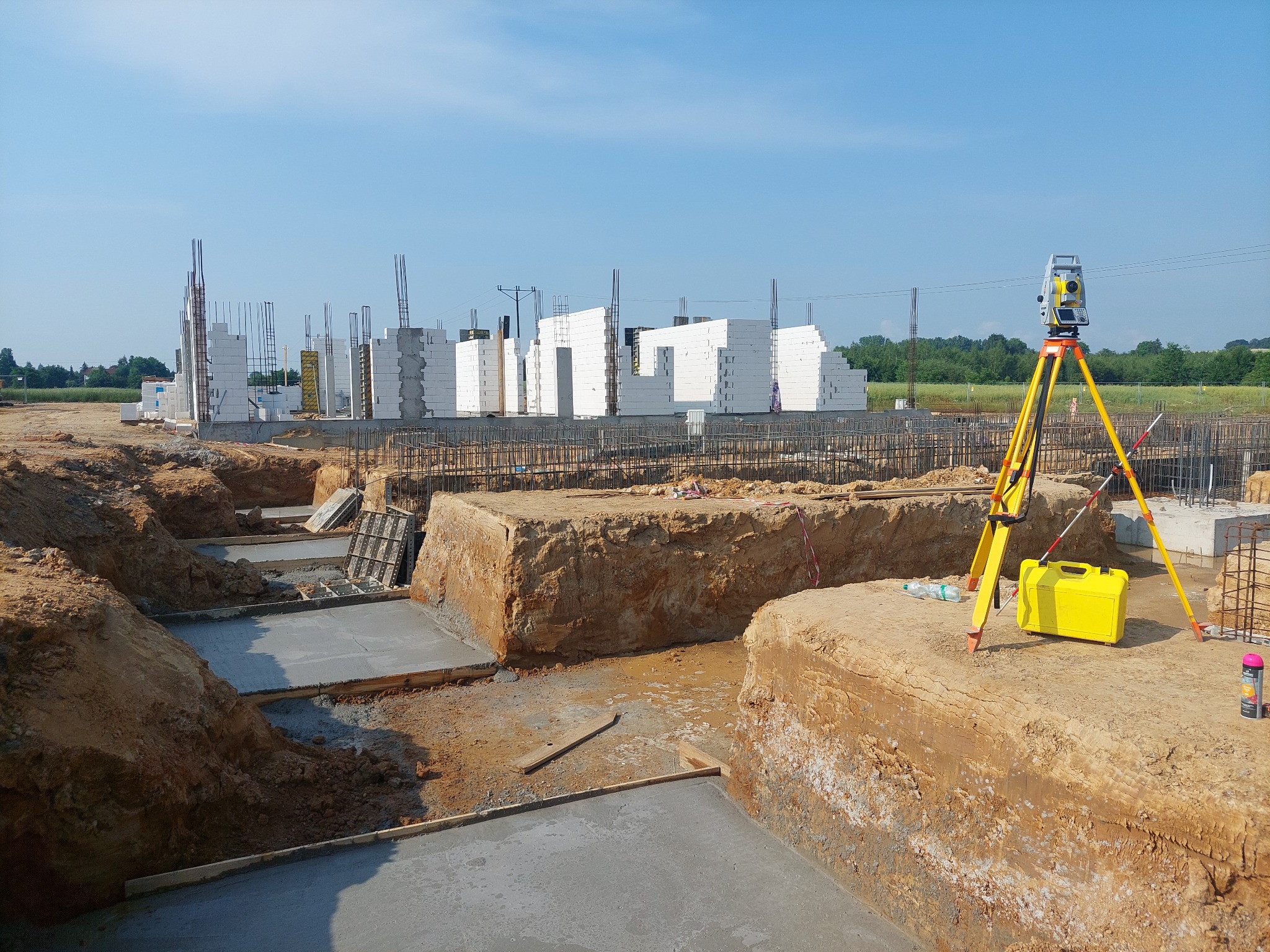 Prace ziemne pod budowę domu jednorodzinnego: wykopane fundamenty, wylany beton, zbrojenia, białe bloczki konstrukcyjne i żółty teodolit na trójnogu.