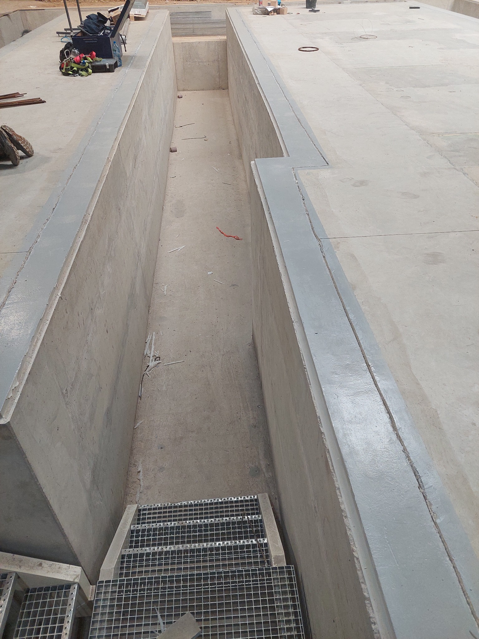 Betonowy kanał inspekcyjny z metalowymi schodami wejściowymi, widok z góry, narzędzia i sprzęt roboczy rozłożone w tle.