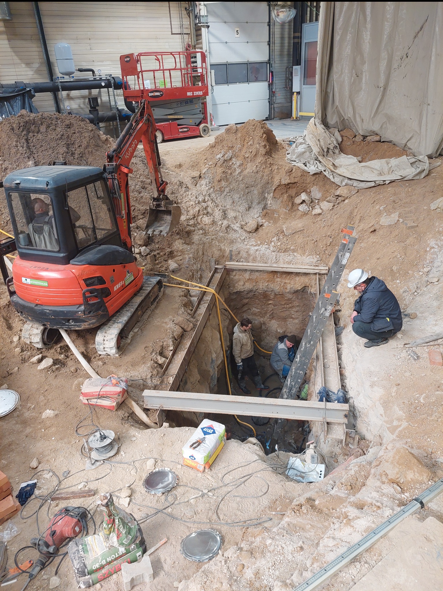 Wykop zabezpieczony stalowymi belkami, wewnątrz pracują trzy osoby, na zewnątrz minikoparka Kubota, obok leżą worki z betonem i narzędzia, w tle podnośnik nożycowy.