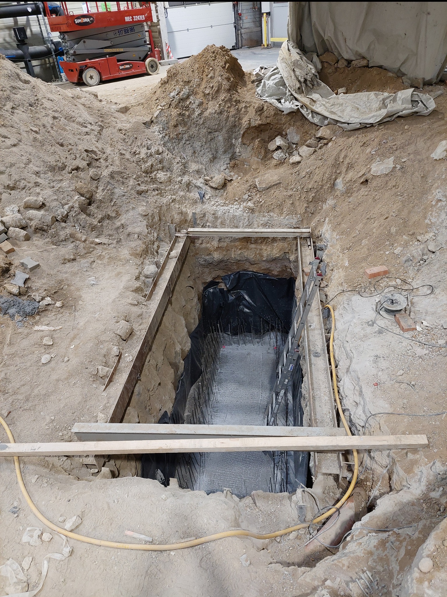 Wykop fundamentowy z deskowaniem i zabezpieczeniem folią, widoczna drabina i podnośnik koszowy w tle.
