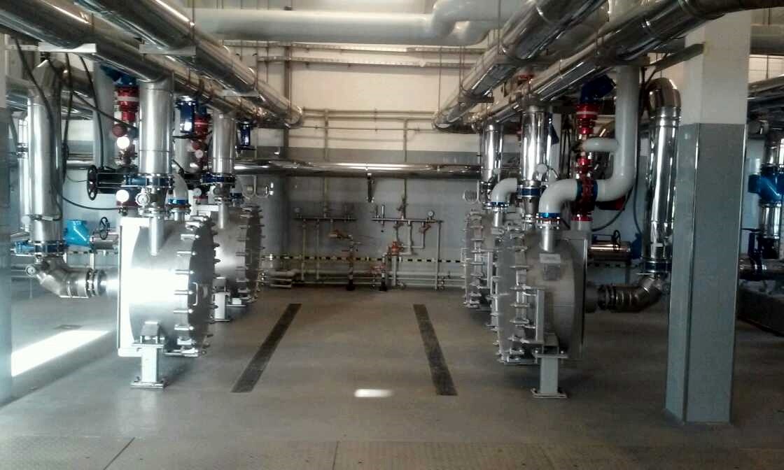 Wnętrze hali z zamontowanymi, srebrnymi instalacjami przemysłowymi: rury, zawory i zbiorniki z licznymi śrubami, ustawione w rzędach.