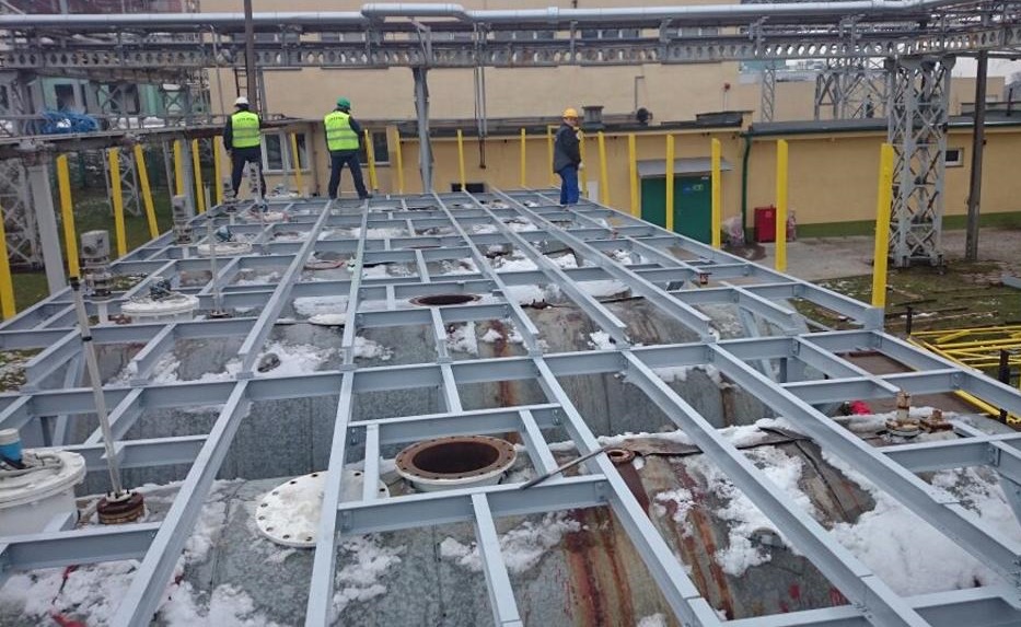 Montaż stalowej konstrukcji kratownicowej na zbiorniku, pracownicy w odblaskowych kamizelkach nadzorują prace.