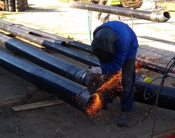 Pracownik w niebieskim kombinezonie szlifuje spaw na czarnej rurze, iskry rozbryzgują się, w tle inne rury na drewnianych podporach.