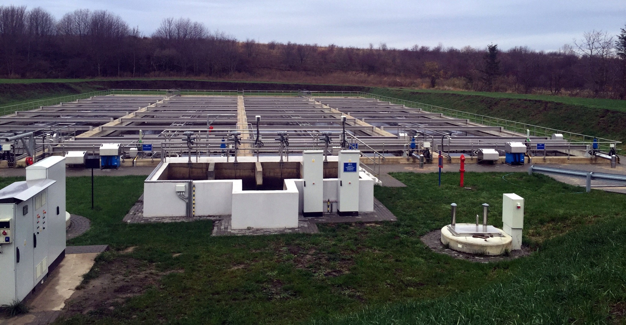 Rozległa oczyszczalnia ścieków z metalową konstrukcją filtracyjną, białymi panelami sterowania i zielonym trawnikiem w tle, widoczne wzgórza z drzewami.