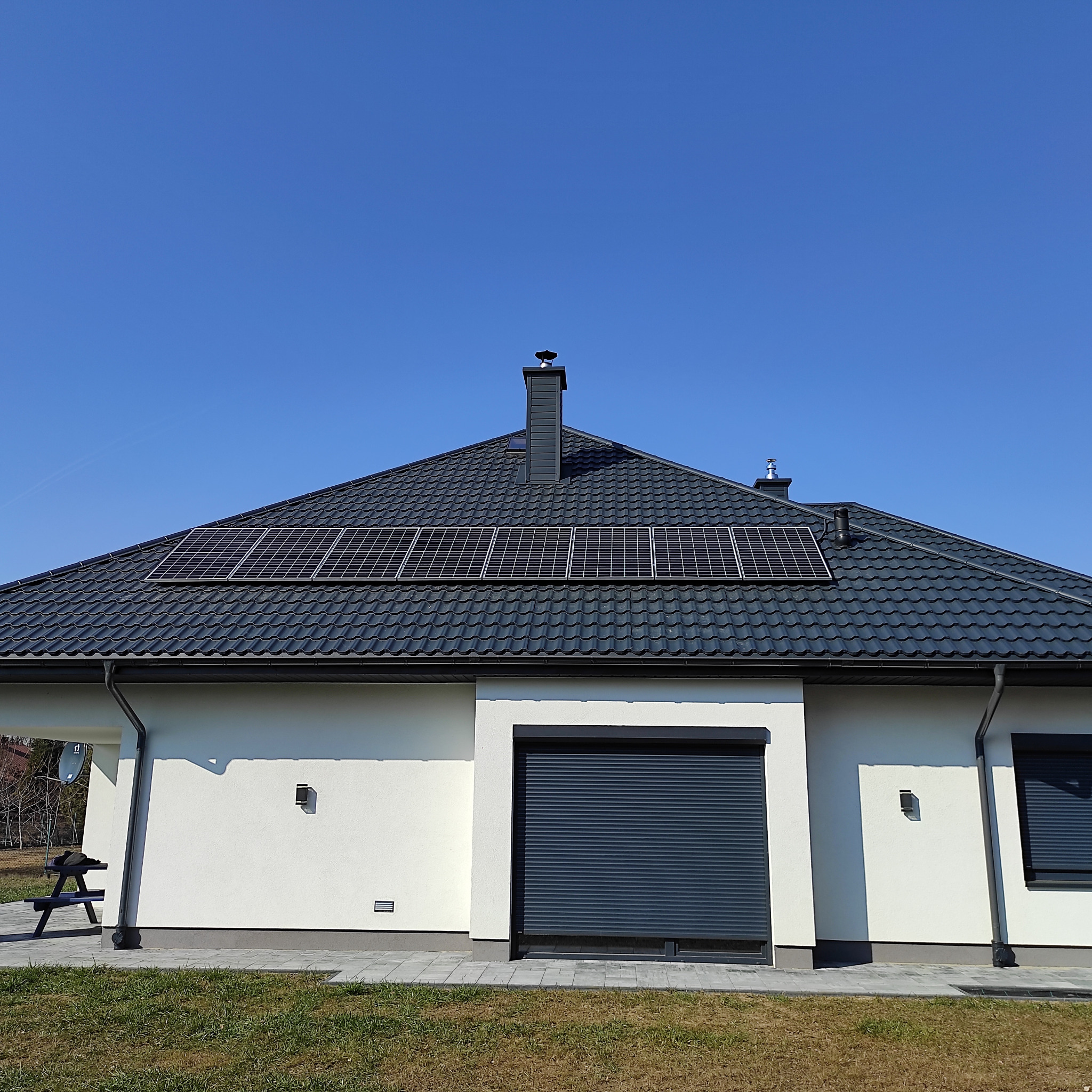 Dom jednorodzinny z ciemną dachówką i panelami słonecznymi na dachu, widok z poziomu trawnika w słoneczny dzień.