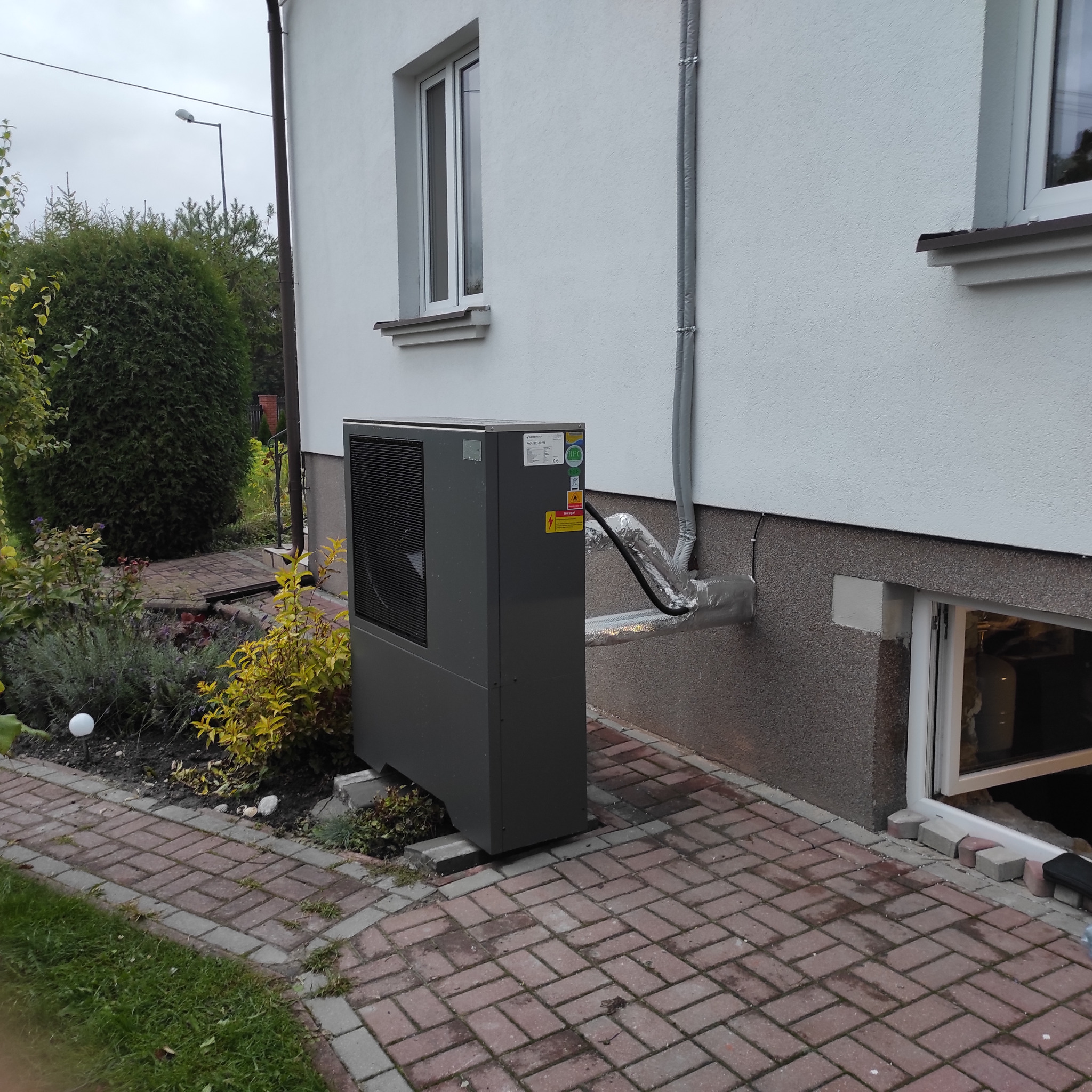 Szara pompa ciepła powietrze-woda zainstalowana przy ścianie domu z oknem, z widocznymi izolowanymi rurami doprowadzającymi, na tle brukowanej ścieżki i roślinności ogrodowej.